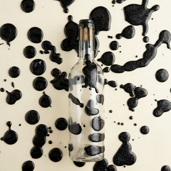 Bouteille en verre ciré de cire dure noir luisante et pailletté avec des taches de cire noire paillettée en fond. 