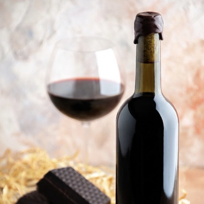 En premier plan il y a une bouteille de vin cirée avec de la cire couleur marron chocolat et en fond il y a un verre de vin rouge avec du chocolat au pied du verre et du foin. 