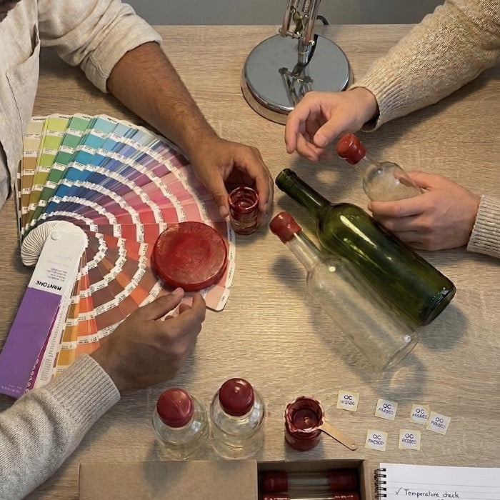 Un duo autour d'une table qui travail avec un nuancier et de la cire ainsi que des bouteilles sur cette table. 